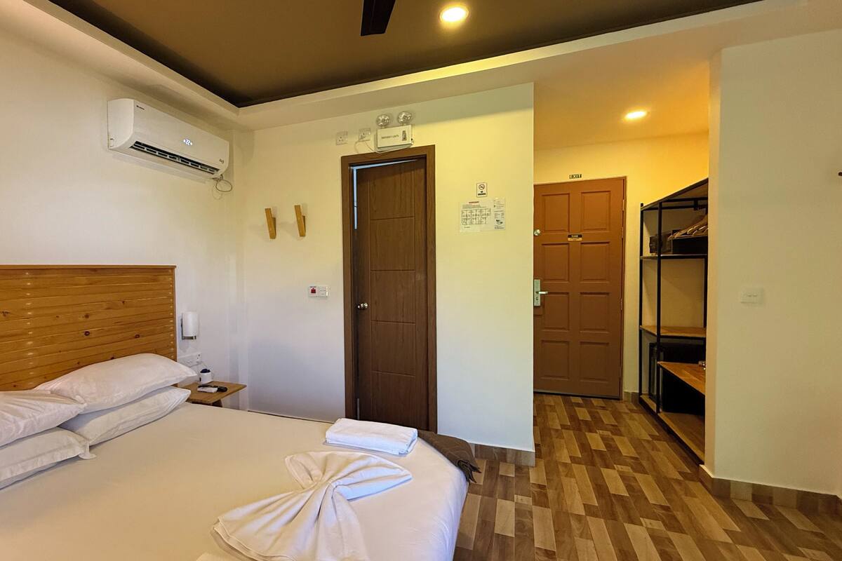 A hotel room with a wooden headboard bed, a bedside table with a lamp, an air conditioning unit, a door with a sign, and a hallway leading to a closet.