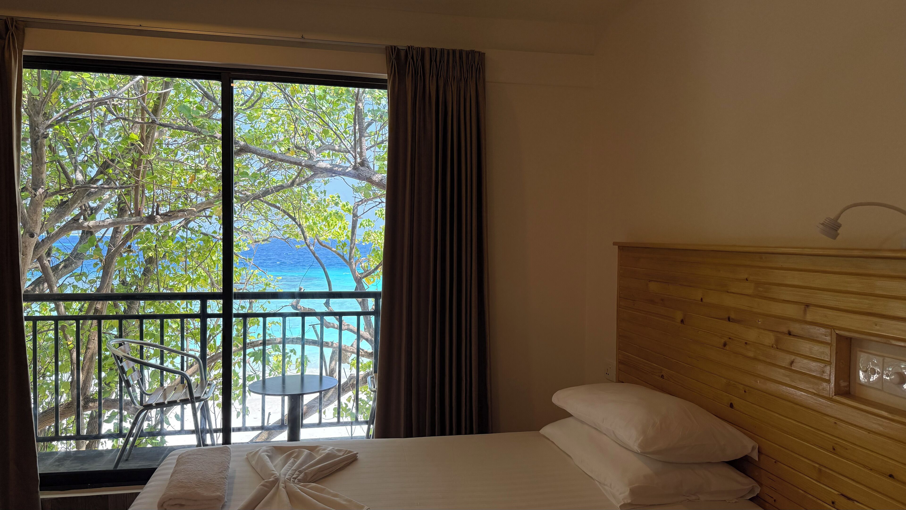 A hotel room with a bed, a wooden headboard, a window with a view of trees and the ocean, and a balcony with a table and chairs.
