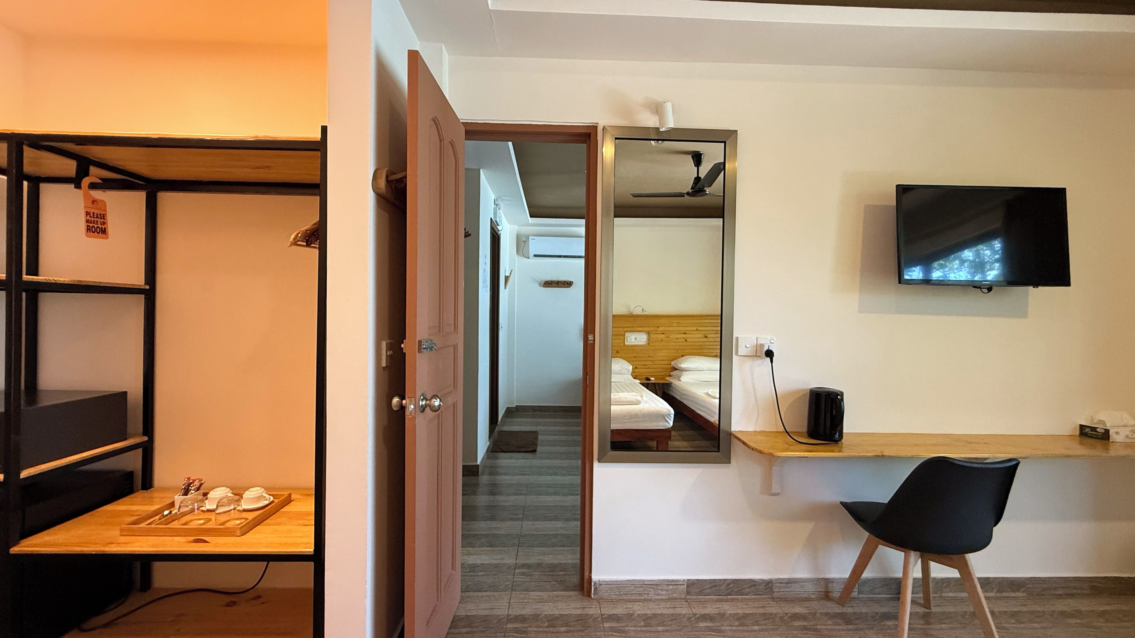 A hotel room with a wooden desk, a black chair, a bed with white bedding, a TV mounted on the wall, and a mirror reflecting the room.