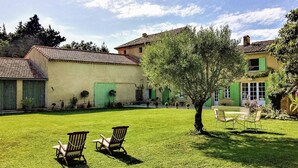 A courtyard with two Adirondack chairs, a small tree, and a building with a tiled roof and green shutters.