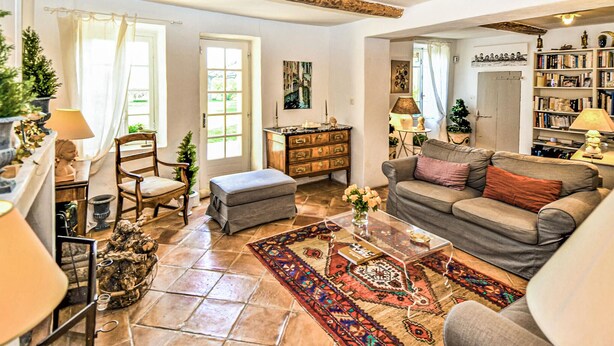 A cozy living room with a sofa, ottoman, and a coffee table. There are two windows with curtains, a fireplace, and a bookshelf filled with books.
