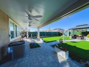 Ein überdachter Patio-Bereich mit Deckenventilator, Gartenmöbel, einem kleinen Tisch und einem dekorativen Windrad.