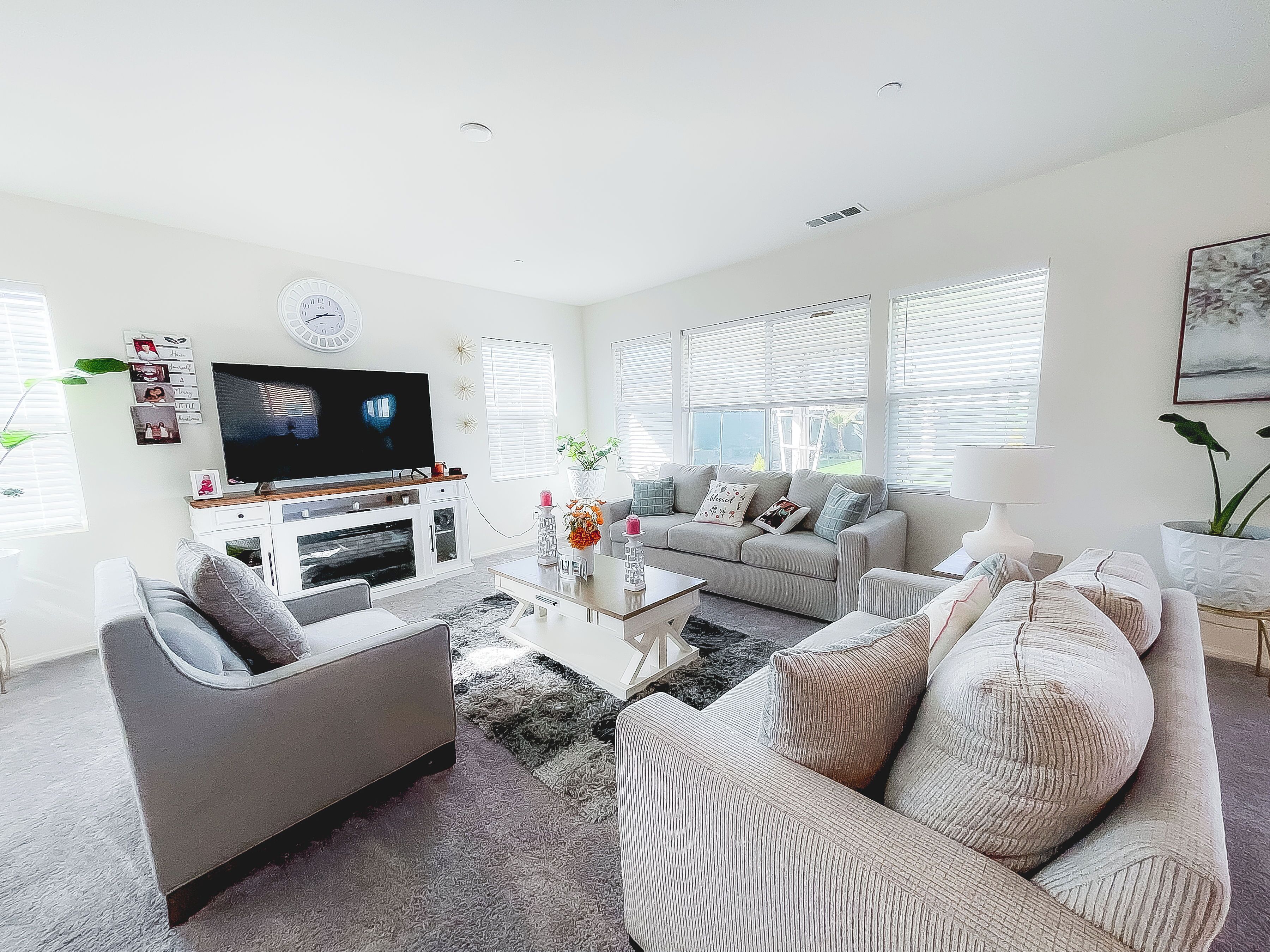 Een moderne woonkamer met een flatscreen-tv, grijze banken, een salontafel en een groot raam met zonwering.