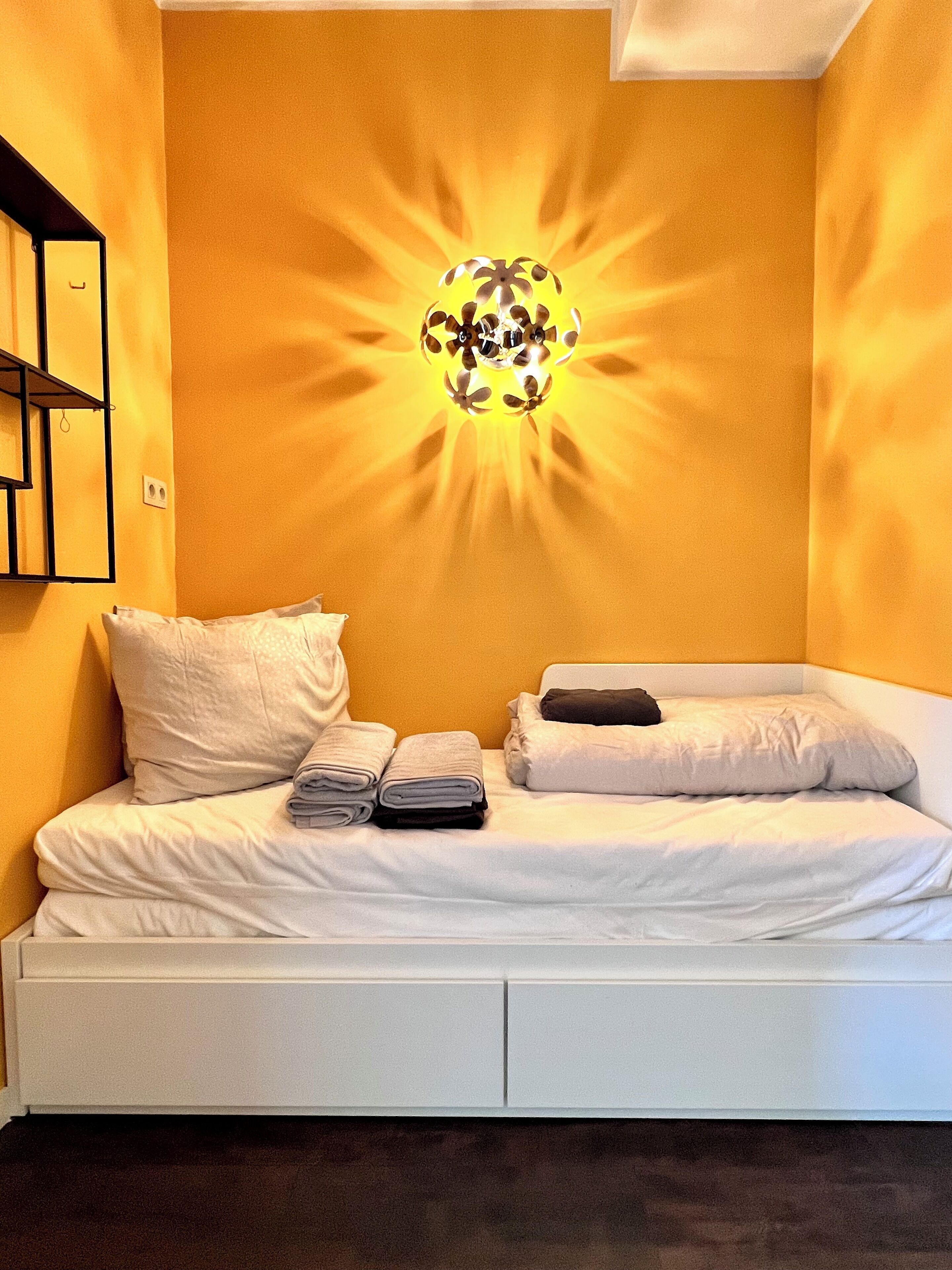 Una cama bien hecha con almohadas y toallas dobladas, una lámpara de pared y un estante con algunos objetos.
