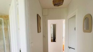 Un pasillo con paredes blancas, suelo de madera y una lámpara colgante. Se observa una puerta a la derecha y una puerta de vidrio a la izquierda.