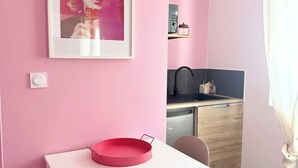A pink-themed room with a white table, a red tray, a framed picture, a kitchen area, and a window with curtains. - Studio rose & spa – Pause bien-être (Quéven)