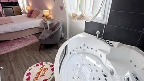 A modern bedroom with a large bathtub, a bed with pink bedding, a small desk with a lamp, and a window with sheer curtains. - Studio rose & spa – Pause bien-être (Quéven)