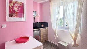 A pink-themed room with a white desk, a chair, a small kitchen area, and a window with sheer curtains. - Studio rose & spa – Pause bien-être (Quéven)