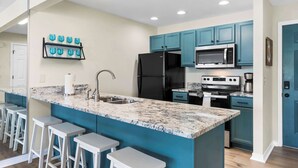 Cocina moderna con armarios verde azulado, un refrigerador negro, microondas y estufa. Hay tres taburetes blancos y una encimera de mármol.