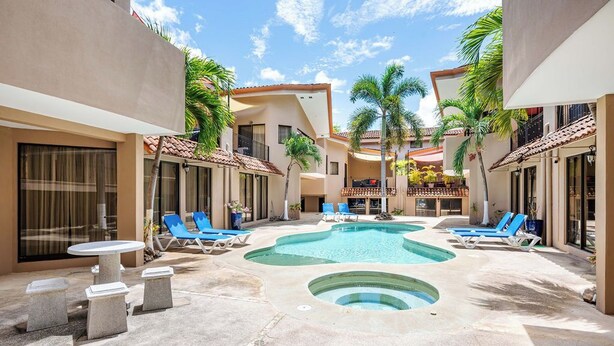 A pool area with lounge chairs and a small table, surrounded by buildings with balconies and palm trees.