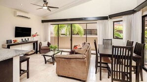 A modern living room with a sofa, coffee table, and dining table. There is a TV mounted on the wall and a ceiling fan.