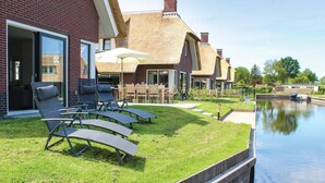 A modern outdoor seating area with lounge chairs and a table, a thatched-roof building, and a canal in the background. - Lovely Home In Idskenhuizen - With Sauna (Idskenhuizen)