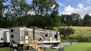 Ein Campingplatz mit Feuerstelle und Stühlen, einem Wohnwagen mit der Aufschrift 'SPORTSMEN' sowie einer grünen Fläche mit Bäumen im Hintergrund.