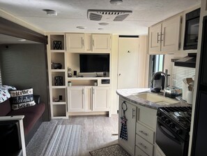 A compact kitchen with a built-in oven, a microwave, and a sink. There is a television on a shelf above the oven.