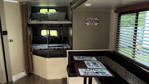 Ein kompaktes Wohnmobil-Interieur mit Etagenbetten, einem Esstisch und einem Spruch ‚Let the adventure begin‘ sowie einem Fenster mit Jalousien.