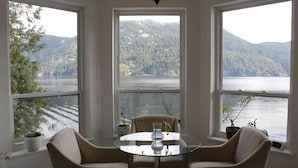 Een kamer met een glazen tafel, twee stoelen en drie ramen met uitzicht op een meer en beboste heuvels.