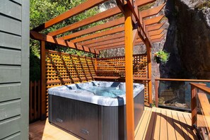A hot tub under a wooden pergola with a wooden deck and a rock wall in the background.