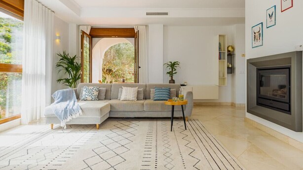 A modern living room with a sectional sofa, a fireplace, a large window with curtains, and a patterned rug.