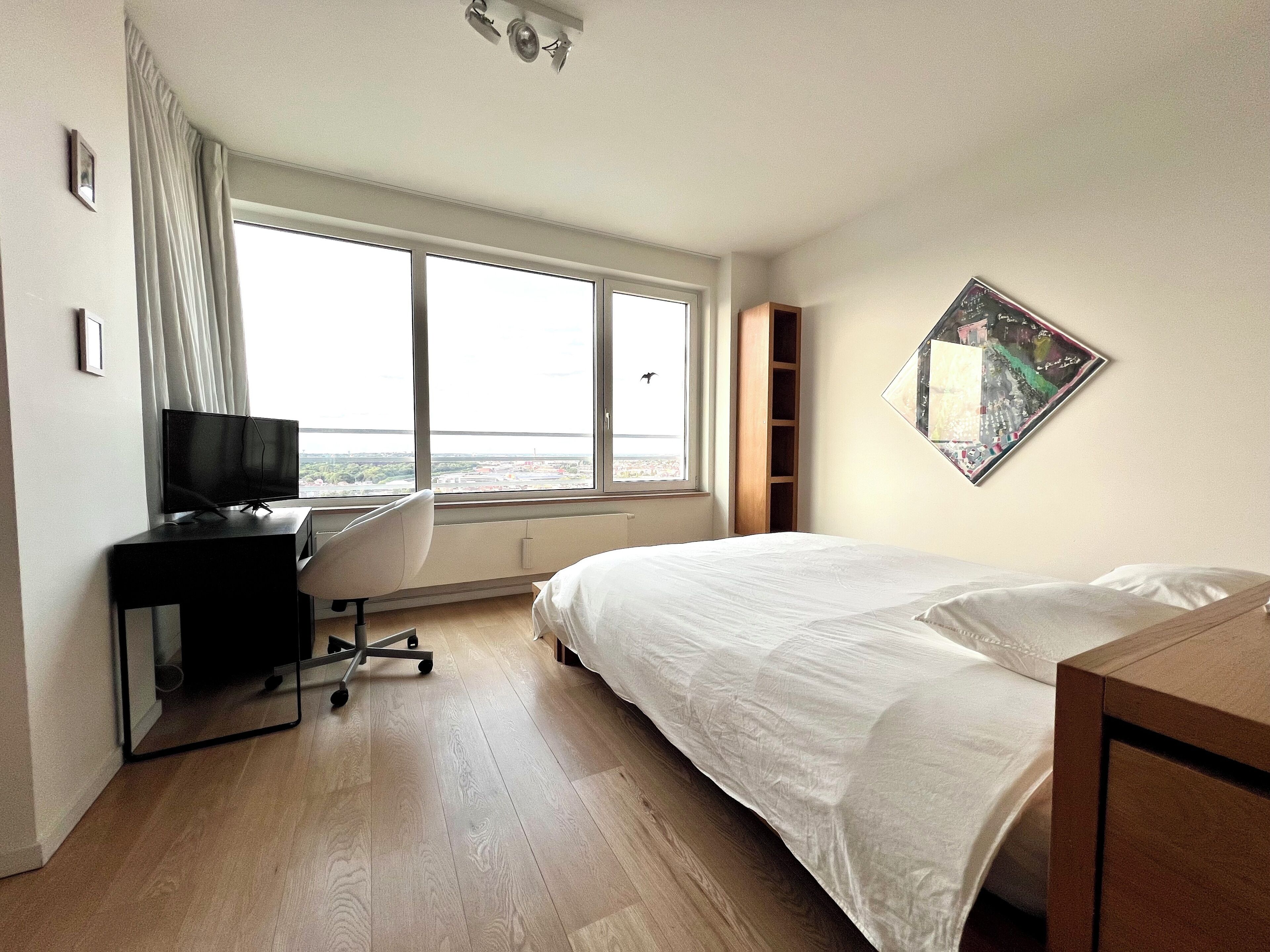 A hotel room with a bed, a desk with a TV, a large window with a view, and a wall-mounted picture.