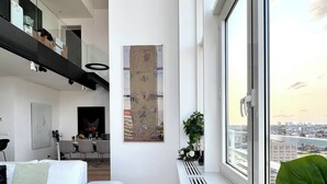 Ein modernes Wohnzimmer mit einem weißen Sofa, einem großen Fenster mit Blick auf die Stadt und einer Küche im Hintergrund.