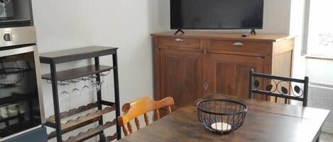 Un comedor con una mesa de madera, sillas, un televisor, un mueble de madera y una zona de cocina con horno y un botellero.