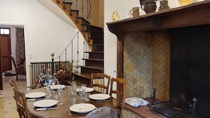 A dining area with a wooden table set for four, surrounded by wooden chairs, a clear plastic chair, and a fireplace with a hexagonal tiled wall. A staircase with a wooden railing leads to another level. - Chez Mamie, Gramat centre (Gramat)