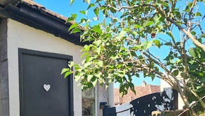 A dark-colored door with a heart-shaped decoration, a window, and a tree with green leaves partially obscuring the view. - Chez Mamie, Gramat centre (Gramat)