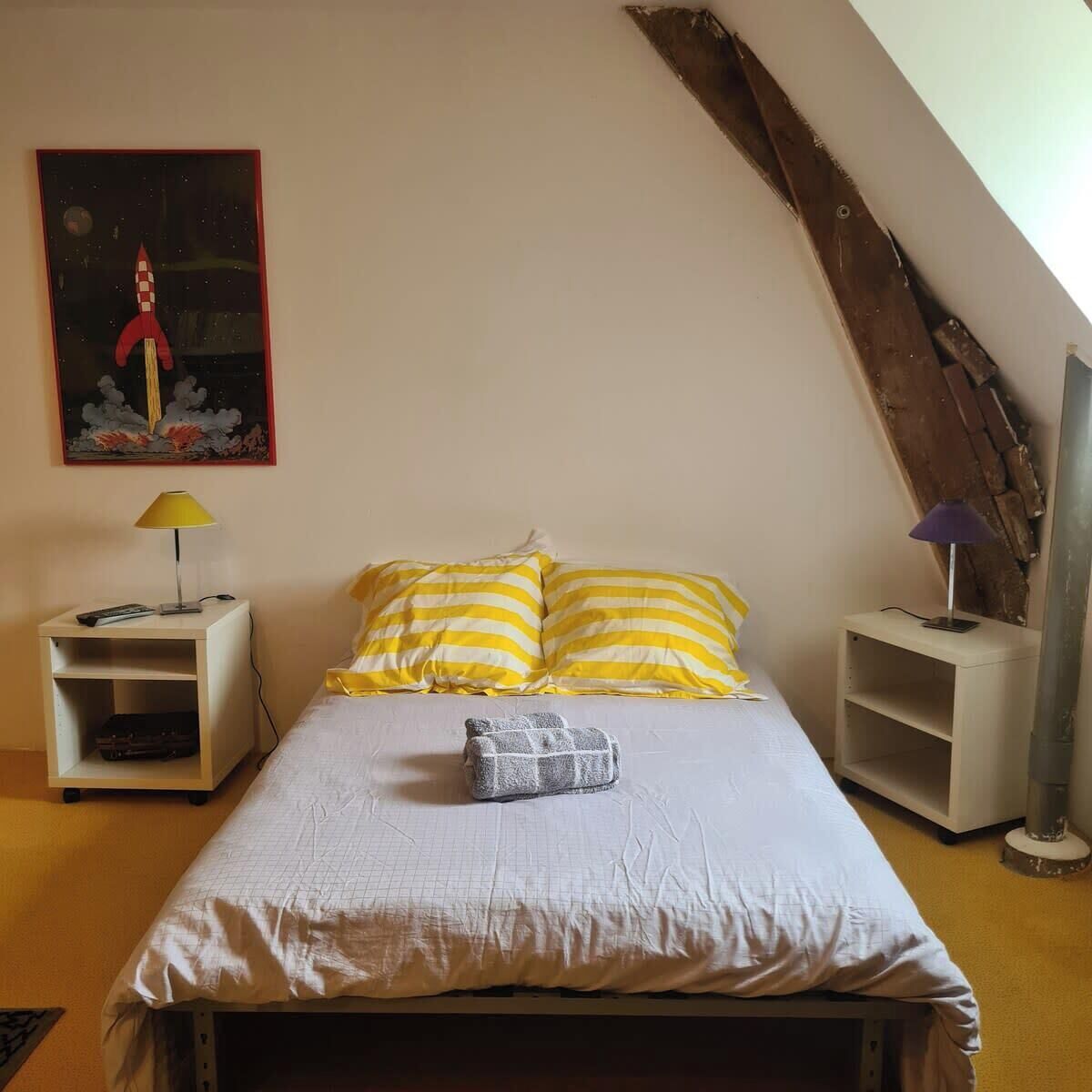 A bedroom with a bed, two nightstands, a lamp on each, a painting on the wall, and a sloped ceiling.