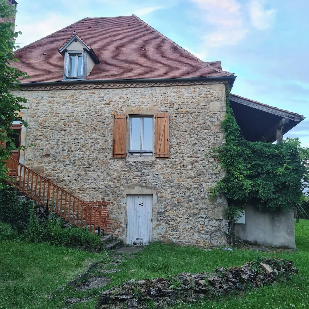 A stone building with a red-tiled roof, a wooden staircase, and a white door.