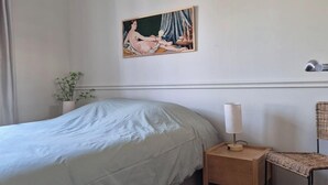 Un dormitorio con cama, una mesita de noche con lámpara, una silla, un cuadro en la pared y una planta.