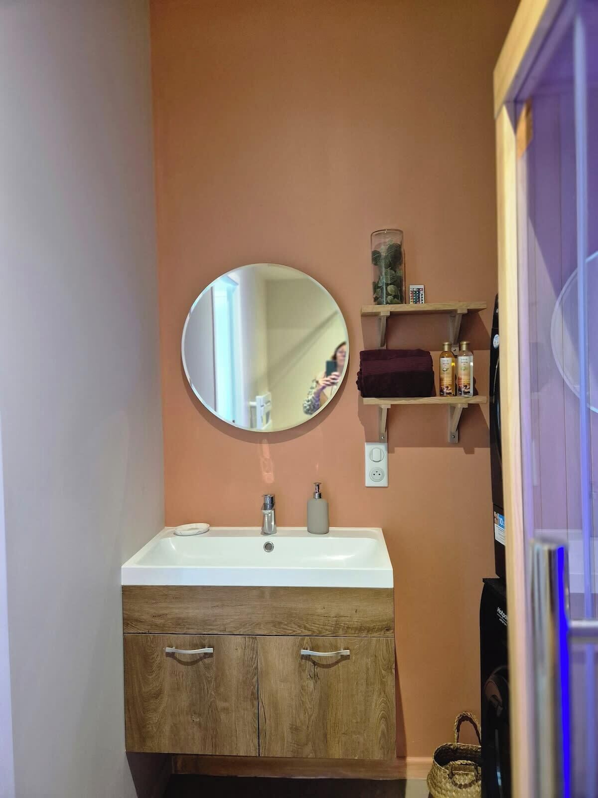 A bathroom with a round mirror, a white sink, wooden cabinets, and a shelf with toiletries.