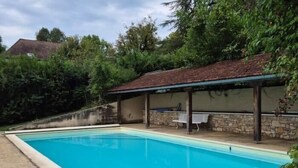 A rectangular swimming pool with a tiled deck, surrounded by a stone wall and vegetation.