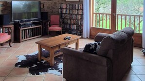 Een gezellige woonkamer met een houten plafond, een televisie, een boekenkast en een groot raam met uitzicht op groen.