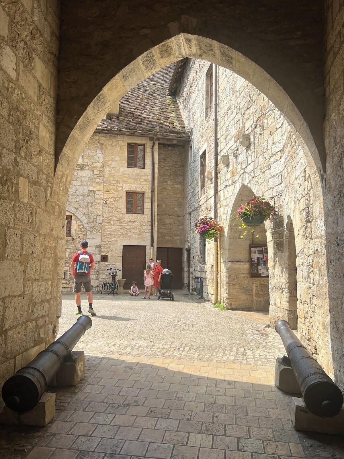 An arched stone corridor with a cobblestone path, leading to a building with windows and a hanging flower basket.
