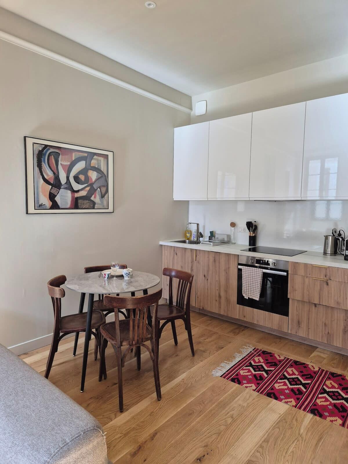 A modern kitchen with wooden cabinets, a dining area with a round table and chairs, and a red patterned rug on the floor.