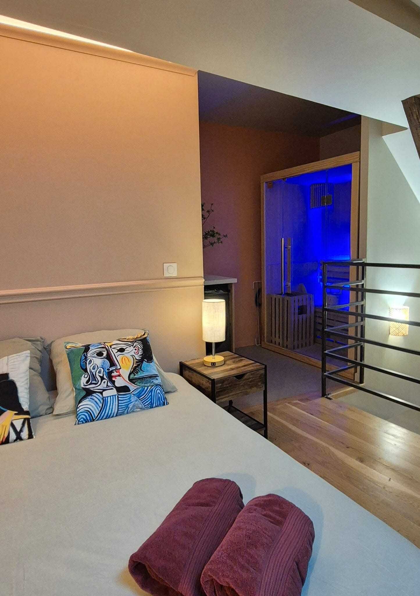 A bedroom with a bed, pillows, a nightstand, a lamp, a wooden floor, and a view of a balcony with a railing.