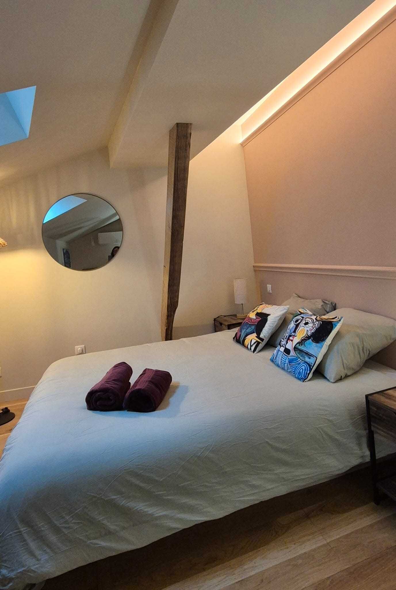 Un lit bien fait, recouvert d’une couette blanche, avec deux oreillers ornés d’un motif de bande dessinée, et une pile de serviettes pliées. Un miroir rond est accroché au mur, au-dessus du lit, et une poutre en bois s’étend parallèlement au lit.