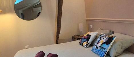 Un lit bien fait, recouvert d’une couette blanche, avec deux oreillers ornés d’un motif de bande dessinée, et une pile de serviettes pliées. Un miroir rond est accroché au mur, au-dessus du lit, et une poutre en bois s’étend parallèlement au lit.