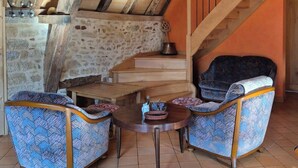 A rustic interior with exposed wooden beams, a stone wall, a wooden staircase, and patterned armchairs around a small round table.