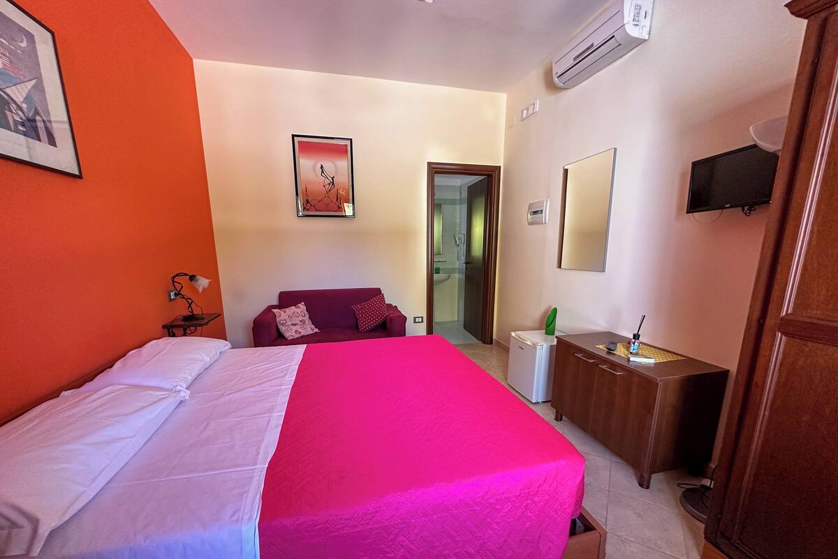 A hotel room with a bed, a desk, a chair, a TV, and a door leading to a bathroom.