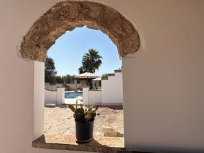 Blick durch ein Rundbogenfenster auf einen Poolbereich, im Vordergrund eine Kakteepflanze.