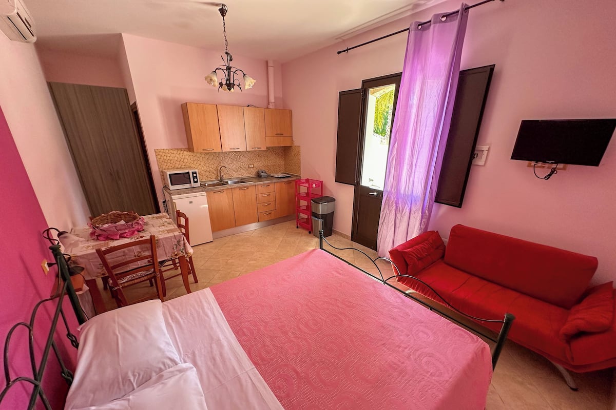 A bedroom with a bed, a red sofa, a kitchen area with cabinets and a microwave, and a television on the wall.
