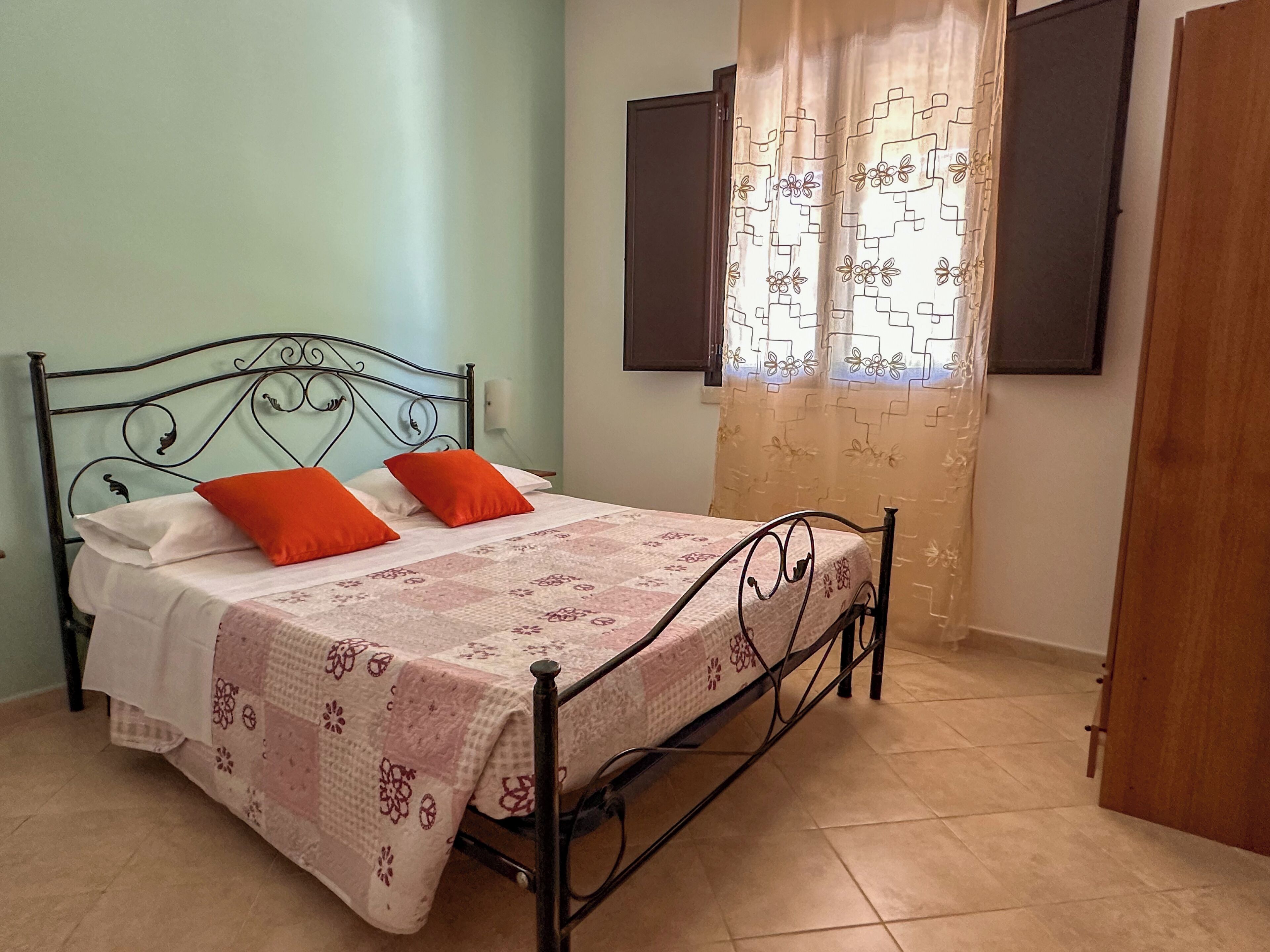 A single bed with a metal frame, two pillows, a patterned bedspread, and a window with curtains.