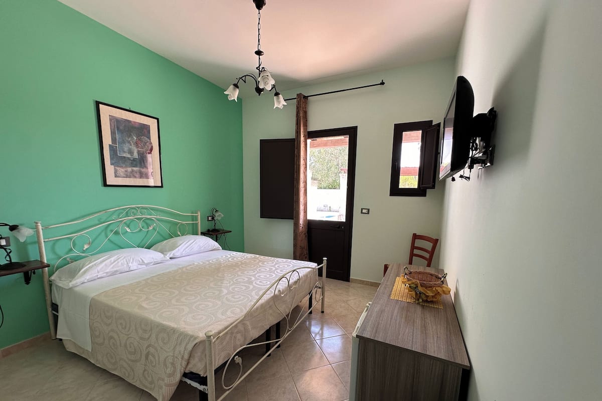 A bedroom with a bed, a bedside table, a chair, a desk, a hanging light fixture, and a window with curtains.