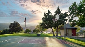 Una cancha de baloncesto al aire libre con un aro, rodeada de árboles y un pequeño edificio.