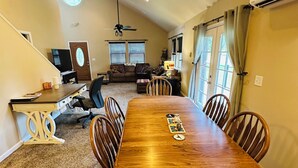 A spacious living room with a large wooden dining table, comfortable seating, a television, and a ceiling fan.