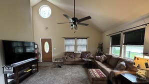 A living room with a TV, sofa, ottoman, ceiling fan, and windows.