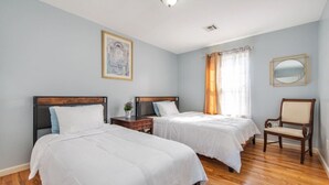 Ein Hotelzimmer mit zwei Betten, Holzboden, einem Sessel, einem Nachttisch, einer Lampe, einem Gemälde und einem Fenster mit Vorhängen.