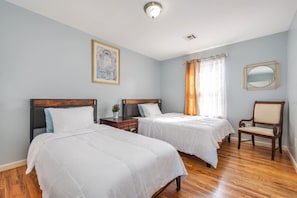 Een hotelkamer met twee bedden, een houten vloer, een stoel, een nachtkastje, een lamp, een schilderij en een raam met gordijnen.
