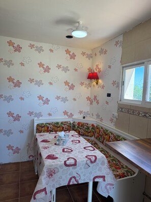 Een kamer met een hoekopstelling, een tafel, een bankje, een plafondventilator en een raam.